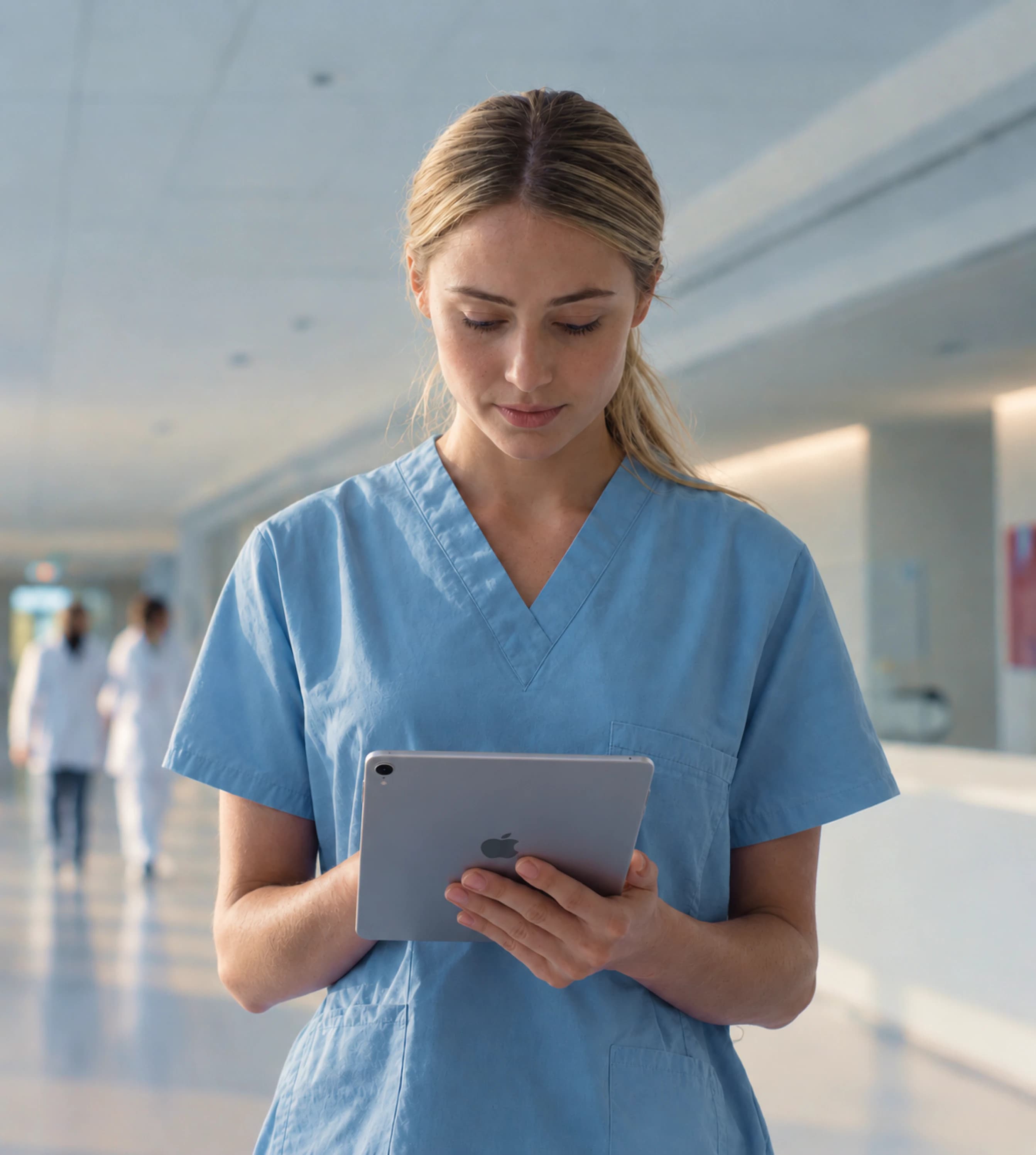 A nurse in scrubs reading a tablet in a hospital corridor